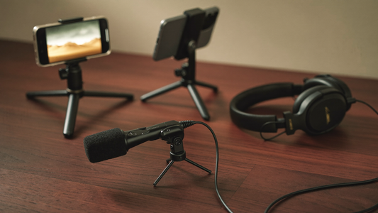Content creator audio setup on a wooden desk with a clip-on lavalier microphone, a phone on a tripod, and over-ear monitoring headphones in soft cinematic light