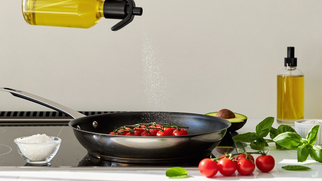 A fine mist of olive oil being sprayed over a pan of cherry tomatoes and basil on a stovetop, with fresh ingredients and a sleek oil dispenser bottle on a bright marble kitchen counter