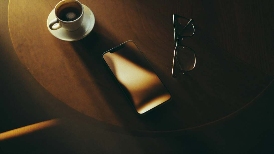 Black smartphone glowing on a dark walnut cafe table beside an espresso and reading glasses, screen content unreadable, illustrating phone screen privacy in public