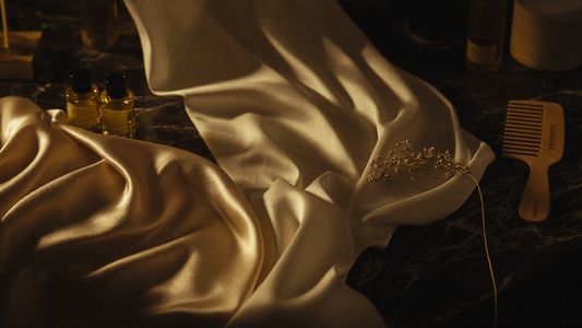 Folds of champagne satin and ivory silk fabric on dark marble beside hair oil bottles and a wide-tooth comb